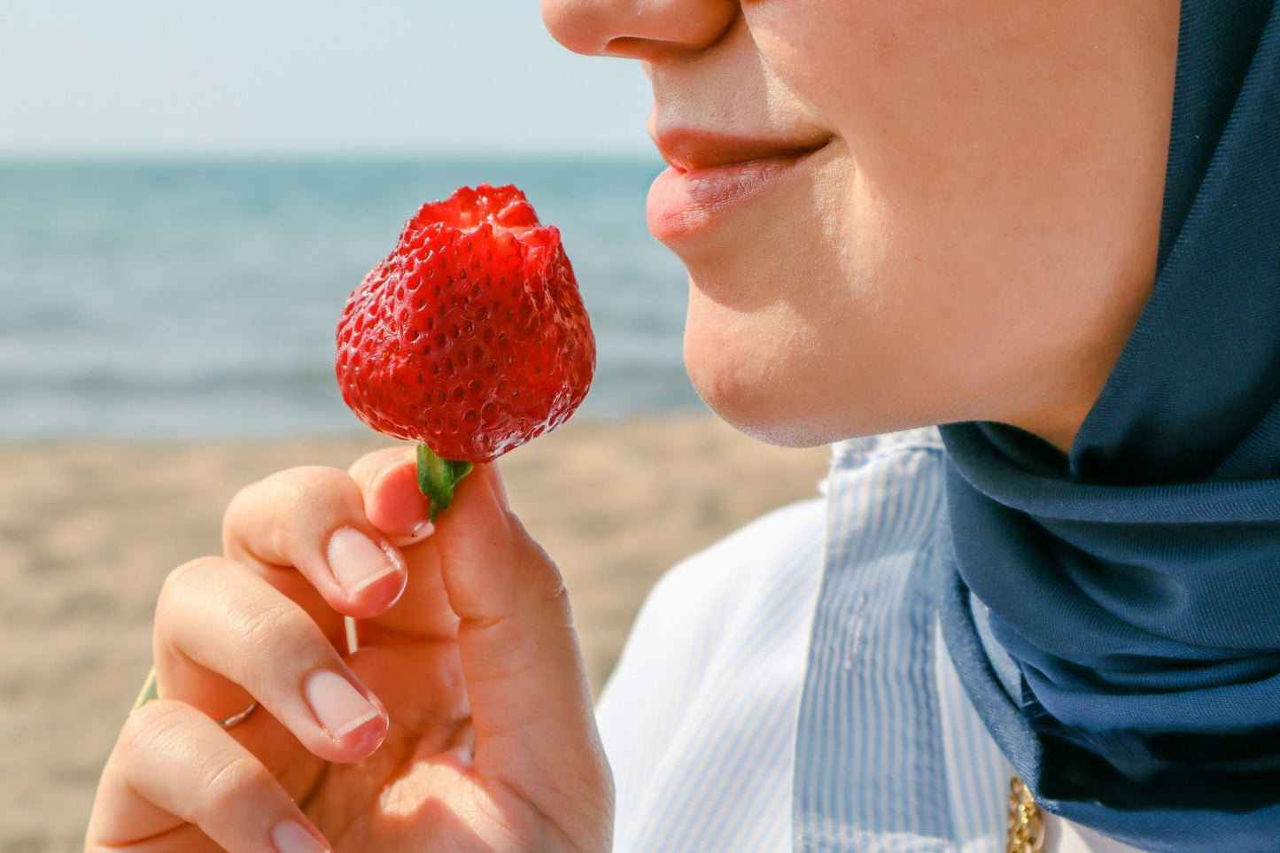A woman in a headscarf enjoys the natural freshness of a strawberry at the beach, highlighting the benefits of using gentle, skin-friendly deodorant for a confident and odor-free day.
